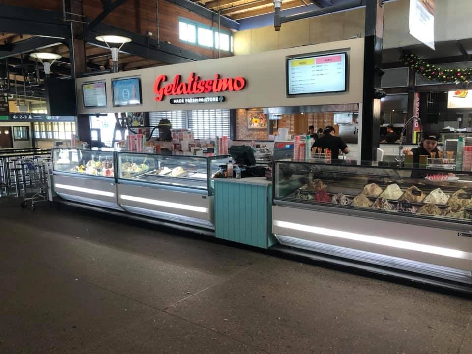 Gelatissimo retail kiosk in a railway station concourse, with illuminated display cabinetry, overhead menu screens and a brick-clad servery counter
